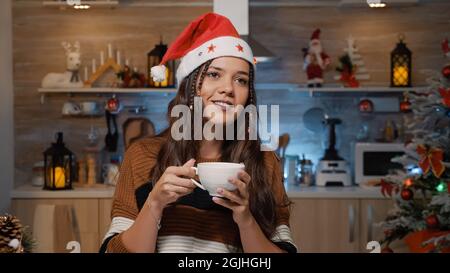 Bonne femme avec chapeau de père noël pensant à l'heure de noël tout en tenant une tasse de thé dans les mains à la maison de fête conçue. Jeune personne se préparant pour un dîner de vacances et des cadeaux pour la famille Banque D'Images