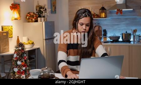 Femme caucasienne assise à la table de cuisine avec ordinateur portable attendant que les invités arrivent pour le dîner de noël dans la maison décorée de fête. Triste adulte vérifiant l'horloge pour des amis et des parents Banque D'Images