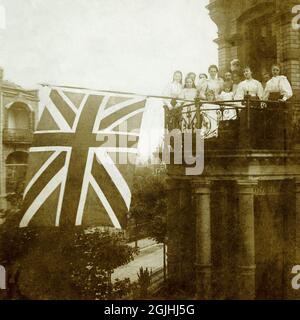 Le 22 juin 1897, le jour du jubilé de diamant de la reine Victoria, jeunes femmes britanniques habillées, élèves d’une école de finition de style suisse à Wiesbaden, en Allemagne, célèbre le 60e anniversaire de son accession au trône du Royaume-Uni en volant d’un grand drapeau d’Union ou d’un drapeau d’Union Jack depuis un balcon. Banque D'Images