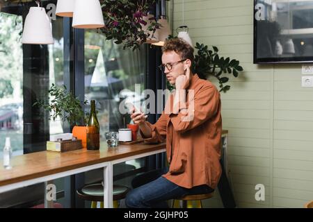 jeune homme dans des lunettes en ajustant les écouteurs sans fil et en utilisant le smartphone dans un café Banque D'Images