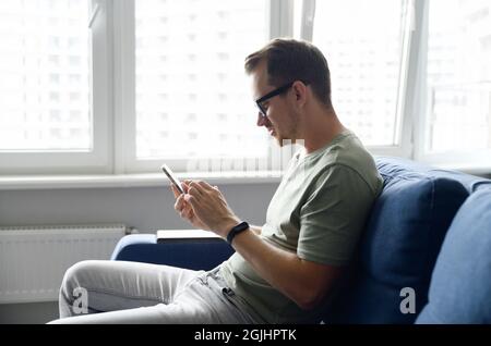 Homme souriant portant des messages de tenue décontractés, envoyant des SMS sur le téléphone intelligent. Vue latérale un jeune homme s'assoit sur le canapé moderne et utilise l'application mobile, les actualités défilant, la navigation sur le Web, le jeu Banque D'Images