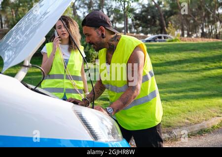 Jeune couple tatoué dans un gilet de sécurité réfléchissant appelant à l'assistance routière. Banque D'Images