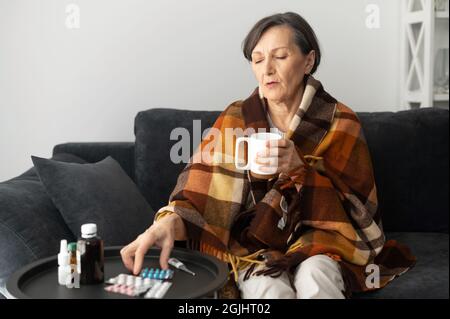 La femme âgée a le froid et la grippe, couverture avec une couverture, s'assoit sur le canapé et boit le remède chaud ou le thé, une femme plus âgée est traitée à la maison, des pilules et des médicaments sur la table près. Concept de propagation de virus Banque D'Images