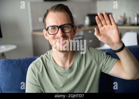 Portrait en gros plan d'un jeune boxeur intelligent à barbe regardant la caméra, portant des lunettes, assis sur un canapé dans le salon et en agitant la main. Le concept de la vue pendant les appels vidéo Banque D'Images
