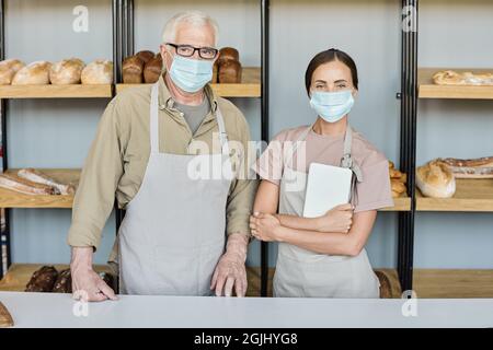 Portrait de boulangers réussis en tabliers et masques debout près du lieu de travail contre présentation avec assortiment de pains Banque D'Images