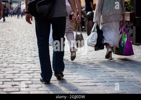 Vue arrière photo des personnes marchant dans une rue pavée | Homme en Jean tenant le téléphone portable à la main et deux femmes portant des sacs marchant dans la rue par beau temps Banque D'Images