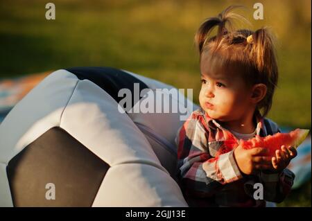 Petite fille mignonne en chemise à carreaux, asseyez-vous sur un pouf de ballon de football et mangez de la pastèque. Banque D'Images
