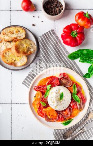 Burrata avec tomates cuites, poivre, oignon rouge et basilic frais sur fond de carreaux blancs. Cuisine italienne traditionnelle. Vue de dessus. Banque D'Images