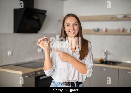 Concept de relocalisation. Une femme excitée se déplaçant dans une nouvelle maison ou un nouvel appartement. Une femme pleine de joie propriétaire regardant la caméra et pointe le doigt sur les clés avec le porte-clés dans sa main Banque D'Images