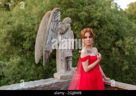 Belle femme tatouée à tête rouge en robe rouge et diadema sur sa tête posant sur le fond flou du château médiéval au coucher du soleil d'été Banque D'Images