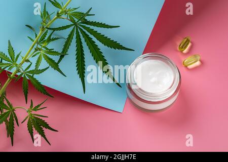 Crème visage de chanvre dans un pot en verre, deux capes d'huile de cbd et feuilles de cannabis sativa vertes sur fond bleu rose. Crème hydratante à la marijuana. Banque D'Images