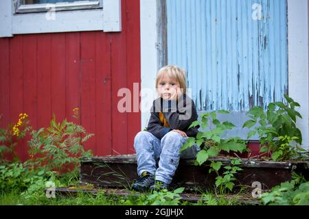 Triste petit enfant, petit garçon d'âge préscolaire, assis sur les escaliers devant la vieille maison ruinée, tristesse Banque D'Images