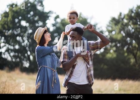 Belle famille multiculturelle de trois vêtus de vêtements décontractés marchant ensemble du terrain d'été. Homme africain tenant un garçon dans les épaules, femme caucasienne les embrassant. Banque D'Images