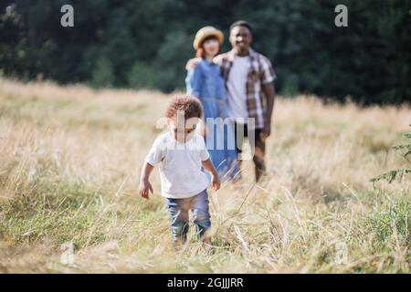 Joli garçon africain courant et jouant dans l'herbe haute tandis que ses parents multiraciaux debout dans des hugs sur fond flou. Concept de famille, bonheur et modes de vie. Banque D'Images