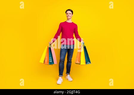 Photo pleine grandeur d'un bon brunet millennium gars stand avec sacs porter chandail rouge jeans isolés sur fond jaune Banque D'Images