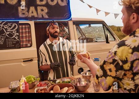 Jeune cuisinier préparant hot dog pour jeune homme pendant qu'ils se tiennent à l'extérieur avec van dans l'arrière-plan Banque D'Images