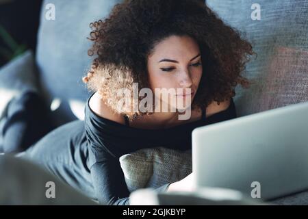 Une jeune femme attirante s'est engrodée en utilisant un ordinateur portable alors qu'elle se détend sur son ventre sur un canapé confortable dans une vue rapprochée Banque D'Images