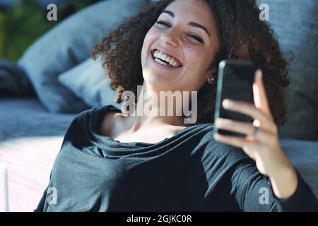 Une jeune femme vivace se chamailler soi-même car elle aime bien rire d'un message ou d'un média sur son téléphone mobile tout en se relaxant à la maison Banque D'Images