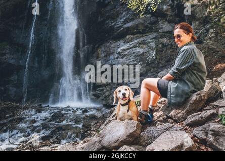 Souriant Femme maître de chien et son ami beagle chien se reposant près de la cascade de la rivière de montagne pendant leur marche ensemble en automne saison. HUMA Banque D'Images