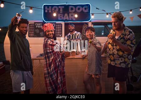 Portrait de jeunes posant à l'appareil photo tout en s'amusant avec de la nourriture et des boissons à la plage fête avec hot dog bar en arrière-plan Banque D'Images