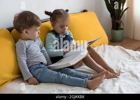 Deux enfants avant-garçonnets lisent un grand livre intéressant de contes de fées sur un lit jaune. Frères et sœurs petits jumeaux lecteur ont plaisir, heureux enfant sur Banque D'Images