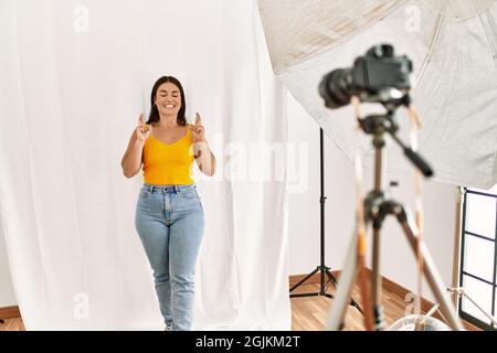 Jeune belle femme hispanique posant comme modèle au studio de photographie gesturant doigt croisé souriant avec espoir et les yeux fermés. Chance et superstititiou Banque D'Images