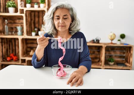 Femme d'âge moyen à cheveux gris mangeant un mètre-ruban assis sur la table à la maison. Banque D'Images