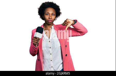 Femme afro-américaine avec cheveux afro tenant le microphone du journaliste avec le visage en colère, signe négatif montrant le désaccord avec les pouces vers le bas, concept de rejet Banque D'Images