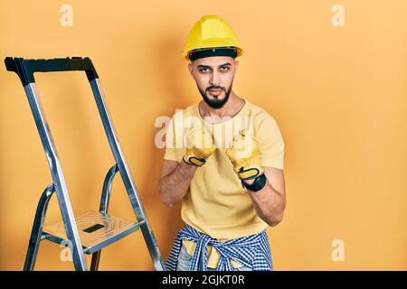 Beau homme avec la barbe par les escaliers de construction portant le casque résistant prêt à combattre avec le geste de défense de poing, en colère et le visage bouleversé, peur du problème Banque D'Images