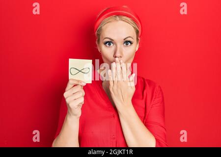 Jeune femme blonde tenant un symbole infini de rappel couvrant la bouche avec la main, choqué et peur de l'erreur. Expression surprise Banque D'Images