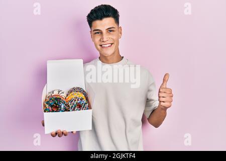 Jeune homme hispanique tenant une boîte de beignets colorés savoureux souriant heureux et positif, pouce vers le haut faisant l'excellent et signe d'approbation Banque D'Images