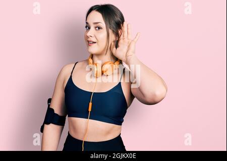 Jeune fille hispanique portant des vêtements de gym et utilisant des écouteurs souriant avec la main sur l'oreille écouter et entendre à la rumeur ou des potins. Concept de surdité. Banque D'Images