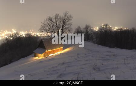 Paysage d'hiver fantastique avec maison en bois lumineux sur fond de lumières de ville dans le brouillard. Chalet confortable dans les montagnes Carpathian. Concept de vacances de Noël Banque D'Images
