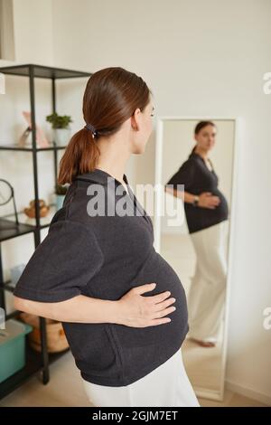 Jeune femme enceinte de race blanche avec queue de cheval touchant le ventre tout en regardant dans le miroir à la maison Banque D'Images