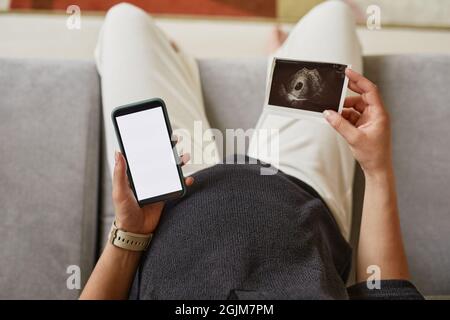 Au-dessus de la vue d'une femme enceinte non reconnaissable utilisant un smartphone tout en envoyant une photo d'image échographique de bébé à son mari Banque D'Images