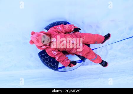 Une fille dans une combinaison d'hiver est assise sur un tubing. L'enfant glisse dans la neige. Banque D'Images