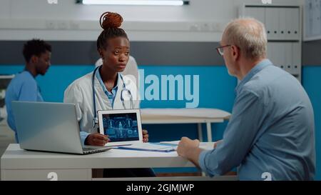Un patient et un médecin expérimenté regardant une tablette moderne avec analyse du corps humain au bureau de l'armoire. Medic ayant un rendez-vous de contrôle avec l'homme aîné tout en utilisant la technologie dans la chambre d'hôpital. Banque D'Images