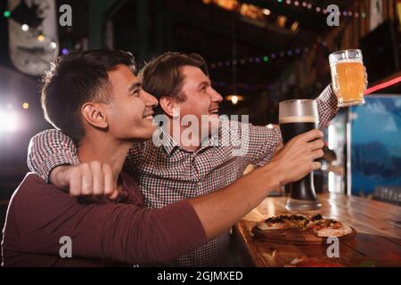 Deux amis de sexe masculin toasting avec leurs verres à bière tout en regardant le match au pub de bière Banque D'Images
