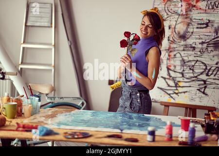 Une jeune artiste féminine tient des fleurs et aime travailler sur une nouvelle peinture dans une atmosphère créative dans le studio. Art, peinture, studio Banque D'Images