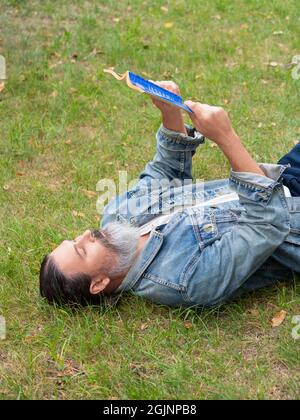 Homme barbu d'âge moyen avec un livre dans le parc. Il est assis sur l'herbe dans le parc et regarde la caméra. Le concept de détox numérique. Banque D'Images