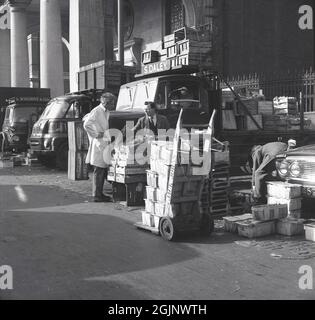 Années 1950, historique, dans la zone de livraison de l'ancien marché de fruits, légumes et fleurs de Covent Garden, dans le centre de Londres, Angleterre, Royaume-Uni, un négociant de marché parlant à un porteur, comme caisses de légumes, y compris le céleri de Hollande, sont déchargés d'un camion sur le terrain de marché de S. Daley. La taille du marché était telle qu'à son apogée, il recevrait près d'un tiers de tous les fruits et légumes importés dans le pays. En raison de la taille dans une petite partie de la ville, le marché a été déplacé en 1974. Banque D'Images