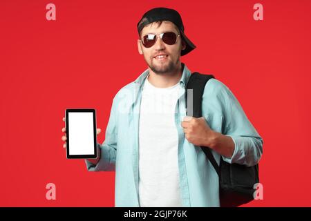 Un touriste européen dans une casquette de baseball et des lunettes de soleil avec un sac à dos sur son épaule se tient sur un fond rouge et montre une tablette. Le concept d'une application mobile pour les voyageurs. Maquette Banque D'Images