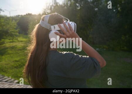 La fille tient des lunettes de réalité virtuelle sur sa tête avec ses mains, se tient contre le fond des arbres verts. Le concept de l'avenir. Photo de haute qualité Banque D'Images