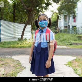 Portrait d'une jeune fille indienne heureuse en uniforme scolaire bleu avec sac rouge et masque de protection allant à l'école Banque D'Images