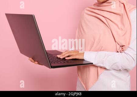 Gros plan portrait d'une femme portant un hijab rose debout sur fond coloré avec un ordinateur portable ouvert dans les mains et en tapant du texte.concept de succès M. Banque D'Images