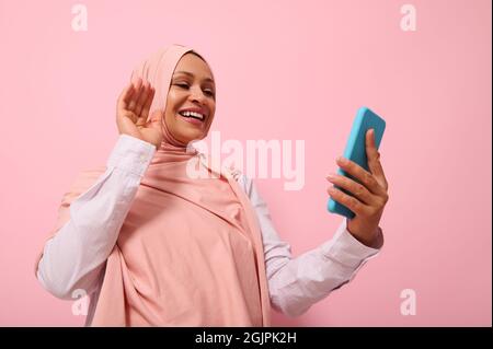 Femme musulmane d'origine arabe ou du Moyen-Orient avec tête couverte de hijab rose ayant une vidéoconférence sur téléphone mobile, saluant son interlocuteur Banque D'Images