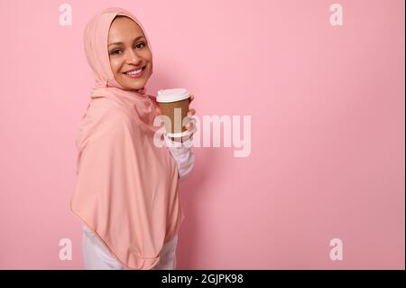 Arabe musulmane gaie femme avec tête couverte dans hijab se dresse trois quarts contre fond rose avec gobelet à emporter de carton jetable de h Banque D'Images
