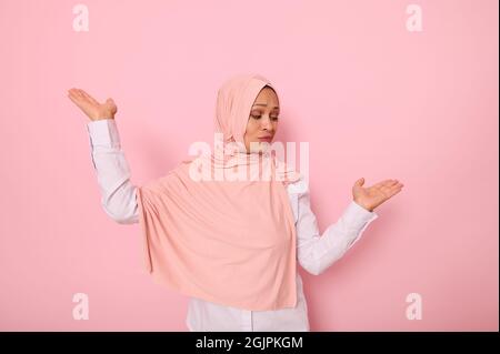 Portrait à la taille d'une mystérieuse femme musulmane portant un foulard hijab, montrant quelque chose sur ses paumes isolées sur un backgrou rose coloré Banque D'Images