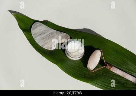 Grattoir de gouache rose, rouleau, pot de crème de feuille verte sur une table blanche. Équipement pour l'auto-massage et les soins de la peau pour le visage et le cou Banque D'Images