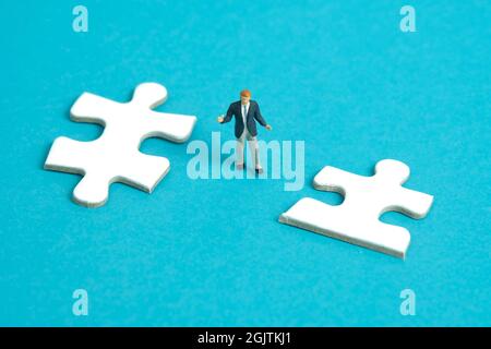 Photographie miniature de personnage de jouet de personnes. Un homme d'affaires haussant debout au milieu de deux pièces de puzzle. Isolé sur fond bleu. PH de l'image Banque D'Images